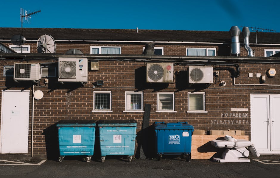 Exterior view of a brick building with seven external air conditioning units and vents mounted along the upper wall, visible beneath a dark sloped roof. Four small windows with white frames are aligned horizontally at the middle of the wall. Below the windows, three large litter bins are placed on the pavement: two blue bins designated for waste disposal and one black bin with white wheels. To the right, a white double door and a sign reading 'NO PARKING DELIVERY AREA' are visible on the building. The scene is illuminated by natural daylight under a clear, blue sky, and the surroundings suggest a residential or commercial property where loading and unloading activities, such as house removals, furniture transport, or packing and moving, occur regularly. Man with Van Palmers Green’s service involves accessing this area for efficient relocation logistics and waste disposal as part of home removals process.
