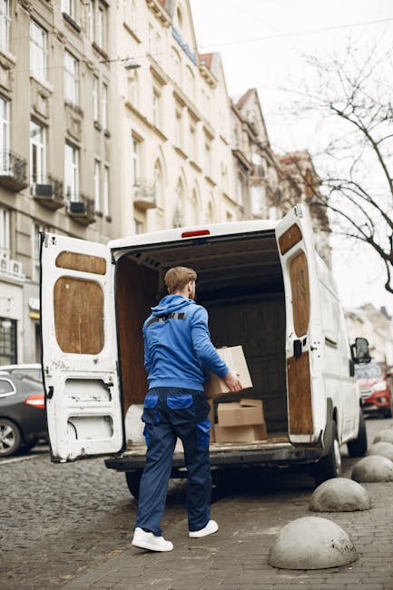 A man wearing a white t-shirt, jeans, and a black cap is transporting three cardboard boxes, stacked one on top of another, towards the open side door of a white panel van parked on a residential stre
