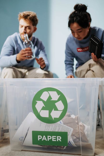 A clear plastic recycling bin with a green recycling symbol and a label reading 'PAPER' is positioned on the floor inside a home during a house removal process. Behind the bin, two movers are visible; one is a man with curly hair drinking from a clear water bottle, and the other is a woman with dark hair tied up, wearing a blue volunteer shirt, crouching and holding a clipboard or tablet. They are in a domestic setting with a light-colored wall in the background, and the scene is lit with soft indoor lighting. The image showcases the process of packing and sorting recyclable materials as part of furniture recycling or home relocation activities, with the movers preparing items for transportation. Man with Van Palmers Green's removal services often include sustainable packing and recycling initiatives, vital for eco-friendly furniture recycling in Palmers Green.