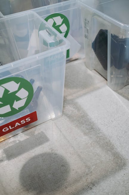 Three transparent plastic recycling containers with green recycling symbols on their sides are positioned on a light-colored, textured floor inside a property. One container has a red label marked 'GLASS' attached to its front, indicating its designated recycling category. The containers are filled with various recyclable items, including pieces of cardboard, plastic, and possibly broken glass or ceramic. The scene shows a clear view of the containers' open tops, revealing the contents for sorting during a home relocation or move. This setup is part of the packing and moving process managed by Man with Van Palmers Green, focusing on eco-friendly furniture recycling efforts in Palmers Green, as part of house removals or furniture transport services.
