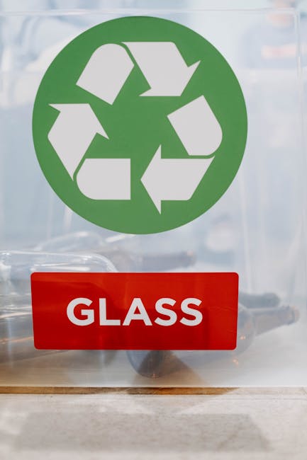 A clear plastic recycling bin with a green recycling symbol and a label reading 'PAPER' is positioned on the floor inside a home during a house removal process. Behind the bin, two movers are visible; one is a man with curly hair drinking from a clear water bottle, and the other is a woman with dark hair tied up, wearing a blue volunteer shirt, crouching and holding a clipboard or tablet. They are in a domestic setting with a light-colored wall in the background, and the scene is lit with soft indoor lighting. The image showcases the process of packing and sorting recyclable materials as part of furniture recycling or home relocation activities, with the movers preparing items for transportation. Man with Van Palmers Green's removal services often include sustainable packing and recycling initiatives, vital for eco-friendly furniture recycling in Palmers Green.