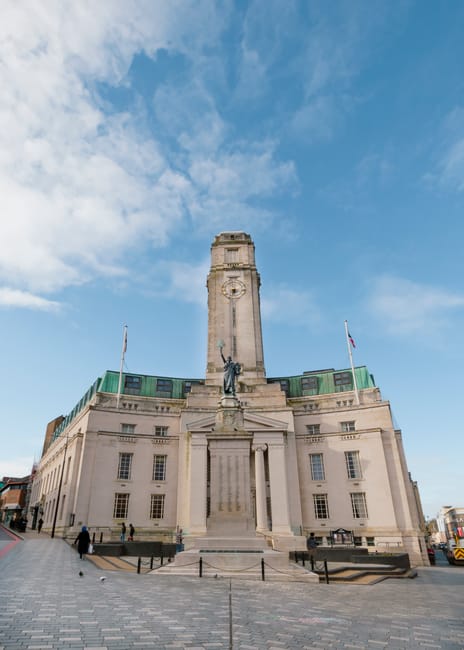 A tall stone monument with a rectangular base and a central clock tower extending upward, situated in an open paved area. In front of the monument, there is a statue of a person holding an object, mounted on a pedestal. Surrounding the monument are a few pedestrians walking along the paved surface. The building has multiple windows and is topped with a green patina roof, with flags flying on either side of the monument. The sky above is mostly clear with some scattered clouds, and sunlight illuminates the scene, highlighting details of the stone structure. This setting is typical for a public square or town centre, where local landmarks and civic monuments are located, and it may be part of a scene depicting a home or office relocation process nearby. Man with Van Palmers Green occasionally conducts removals and furniture transport in this area, supporting efficient moving logistics in the community.