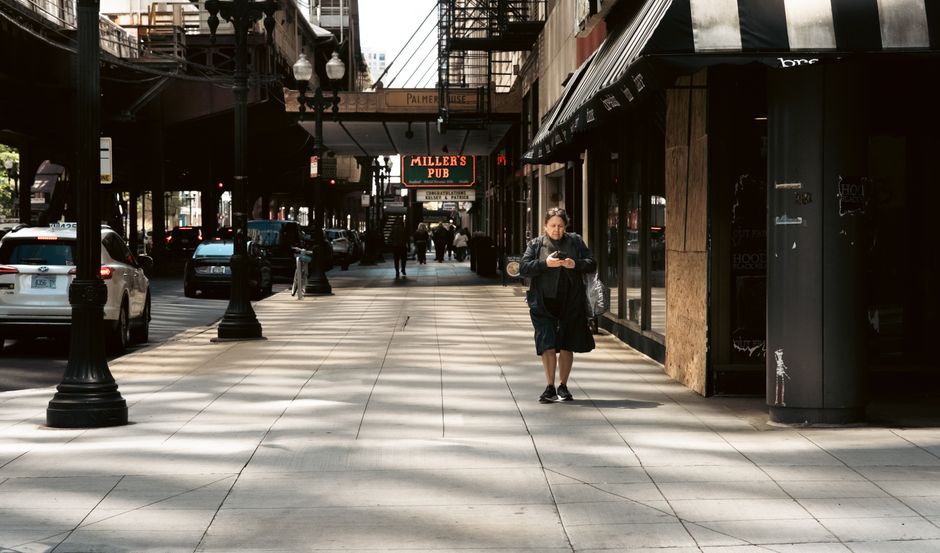 A man with a backpack and black shorts is walking along a wide, paved sidewalk in Palmers Green, with a mobile phone in his hand. On his left, there are black lampposts lining the street, and parked cars including a silver vehicle and a darker one further back. On his right, there is a row of shops with large glass windows and black awnings, with some graffiti visible on a black pillar near the sidewalk. In the background, there are a few pedestrians strolling along, and a distinctive, illuminated pub sign for 'Miller's Pub' hanging over the street. The scene is lit with natural daylight, casting shadows across the pavement, and the area looks busy yet relaxed, representing a typical urban environment where house removals or furniture transport might occur, with Man with Van Palmers Green providing moving and relocation services in this setting.