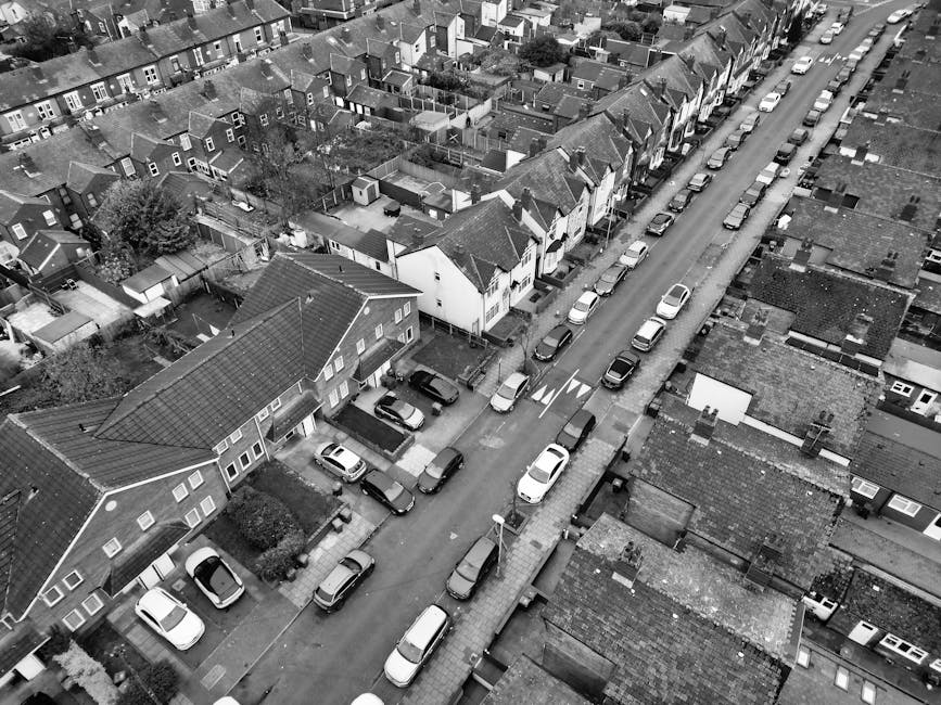 An aerial black-and-white photograph of a residential street showing closely spaced houses with pitched roofs, some with small backyards and garden sheds. Cars are parked along both sides of the narrow road, with a white van positioned near the bottom left corner, possibly engaged in a home relocation or furniture transport activity. The street has a slight turn, and the pavement includes a sidewalk with a streetlamp and a road marking indicating a pedestrian crossing. The tops of the houses display various roofing materials, and the scene is well-lit, highlighting the details of urban home environments. This image reflects the logistical considerations of house removals in residential neighbourhoods, as handled by professionals such as Man with Van Palmers Green, for efficient moving and packing processes.