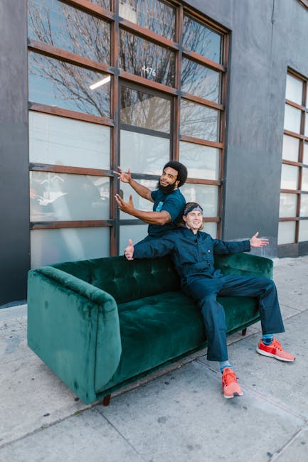 Two men are positioned outdoors, in front of a large windowed building, with one standing behind a green velvet sofa and the other sitting on it. The man standing has short dark hair, a beard, and is wearing a dark blue t-shirt and jeans, smiling while gesturing towards the sofa. The man seated has short, light-colored hair, is dressed in a navy jacket and dark trousers with bright red sneakers, and is extending his arms outward with a cheerful expression. The scene appears to be part of a home relocation or furniture transport process, with the sofa placed on the pavement outside the building, indicating a loading or unloading activity. The environment is well-lit, with natural daylight and a clear sky reflected in the window panes, and the building’s grey exterior wall features large wooden-framed windows, some of which are frosted or partially covered, highlighting a professional approach to moving or packing services offered by Man with Van Palmers Green.
