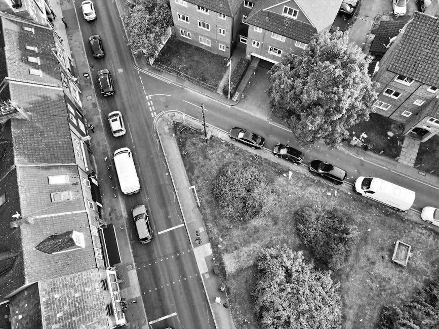 A black-and-white aerial photograph showing a residential street with parked cars lining both sides of the road, including a white van used for home relocation positioned near the pavement. In the foreground, a large van is parked adjacent to an open doorway, possibly for loading furniture and boxes during a move. The street is narrow, with a paved sidewalk on one side and a small grassy area with trees and bushes on the other, indicating a suburban neighbourhood. Nearby, multi-storey apartment buildings with visible windows and balconies are seen, and a few cardboard boxes and packing materials may be present on the pavement or near the vehicles, suggesting active packing or loading during a removal process. This scene aligns with house removals and furniture transport services, supported by the presence of a professional moving van, as well as equipment such as trolleys or moving blankets, typical for a home relocation scenario involving careful packing, loading, and transport.