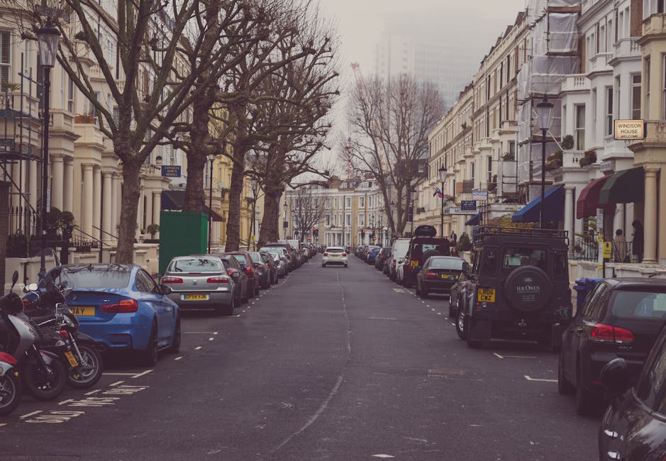A narrow residential street in Palmers Green with parked cars lining both sides of the road, including a blue car and a black SUV in the parking bays, and motorcycles on the pavement. Tall, leafless trees extend overhead, creating a canopy over the street. Victorian-style terraced houses with white facades, bay windows, and decorative railings are visible on both sides, some with awnings and potted plants on the balconies. The street is well-lit by vintage-style lampposts, and a green parking suspension notice is visible on a post near the sidewalk. The pavement is clear, and the overall scene suggests a quiet, urban home relocation area with vehicles ready for furniture transport or packing and moving activities, as managed by Man with Van Palmers Green, with considerations for local parking suspensions for efficient moving logistics.