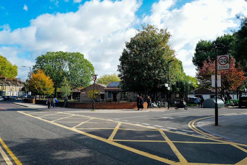 A daytime street scene in Palmers Green showing a paved intersection with yellow box markings and a 20 mph zone sign. In the background, there are trees with green, orange, and red foliage, and residential buildings with brick walls and pitched roofs. Several pedestrians are walking along the pavement, some near bicycles parked beside a black metal fence. A group of movers from Man with Van Palmers Green is visible, loading or unloading furniture and boxes wrapped in plastic and cardboard, using trolleys and equipment near a white van parked on the side of the street. The scene emphasizes the process of home relocation, with items carefully packed and transported, illustrating a typical house removal in a suburban neighbourhood.
