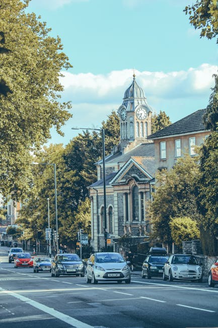 A street scene in Palmers Green under a clear blue sky, featuring a row of parked cars along the pavement with a mix of small family vehicles and sedans, adjacent to a traditional church building with a prominent clock tower and ornate architectural details. Tall trees with green foliage border the street, partially shading the parked vehicles and providing a natural frame around the church. The sidewalk is visible with some pedestrians and street lamps lining the area. This setting illustrates a typical residential and community environment suitable for house removals and home relocation services, such as those offered by Man with Van Palmers Green, highlighting the urban context in which furniture transport, packing, and loading operations can occur efficiently.