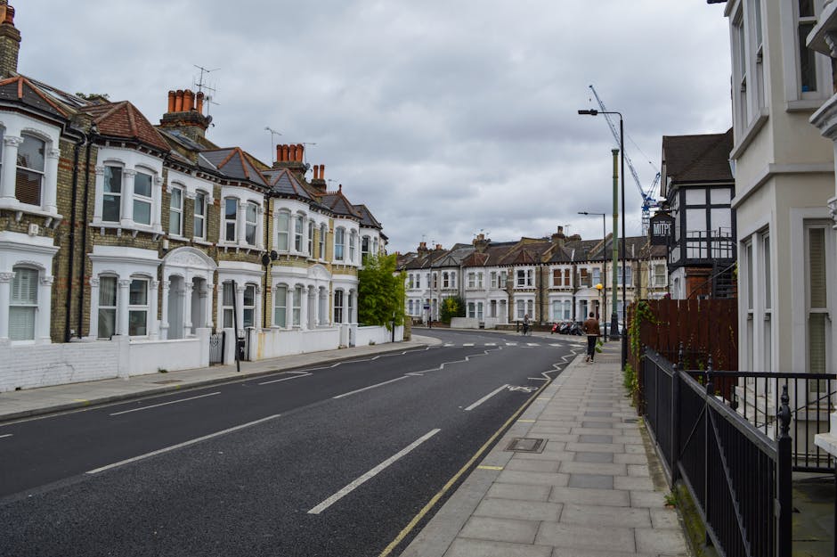 A wide view of a residential street in Palmers Green with a row of Victorian-style terraced houses featuring bay windows, ornate brickwork, and red-tiled roofs. The street is lined with pavement, black metal fences, and a few parked cars. Overhead, cloudy skies cast diffused lighting across the scene. On the right side, a white building with large windows and a small front garden is visible, with a person walking on the pavement. In the distance, a crane can be seen above some construction work, indicating ongoing development. The scene depicts a quiet, established neighbourhood suitable for home relocation and furniture transport services provided by Man with Van Palmers Green, with no active moving activities visible but a setting prepared for efficient house removals and packing processes.