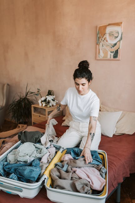 A woman with dark hair tied in a bun, dressed in a white t-shirt and light-colored trousers, sits on a bed in a room with warm-colored walls. She is engaged in packing or organising clothing, which is spread out and folded into two open suitcases placed on the bed. Behind her, there are decorative pillows and a wooden bedside table with a small potted plant and flowers. On the wall, there is a modern abstract painting. The scene captures a home relocation or packing process, with the woman preparing belongings for transit, a task often associated with professional removals services such as those offered by Man with Van Palmers Green.