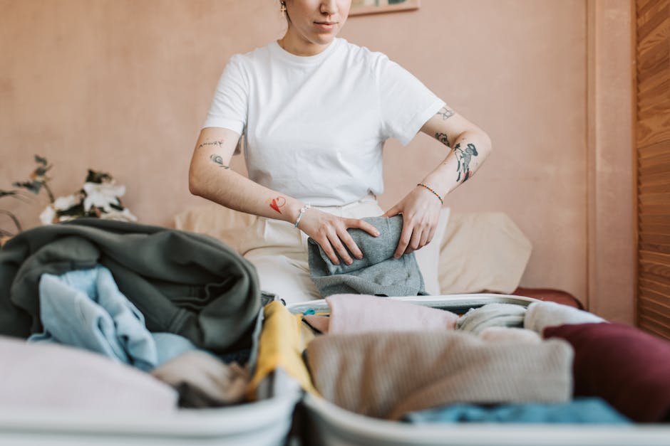 A woman with tattoos on her arms is seen packing clothing into a grey fabric laundry bag, with various other folded clothes and packing materials scattered across a bed. She is wearing a plain white T-shirt and standing inside a home, near a beige wall and a bed with a cream-colored headboard. Visible objects include a partially opened suitcase or packing box containing clothing, a small bouquet of flowers on a table to the side, and a folded blanket or towel. The scene captures the process of home relocation, specifically packing and organising belongings for moving, with natural indoor lighting illuminating the room. This image illustrates the activities involved in a professional removals service, such as those provided by Man with Van Palmers Green, focusing on packing tips for flats on the Palmers Green Triangle, PALMERS GREEN.