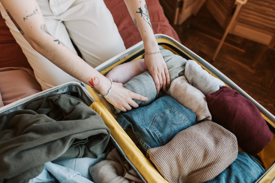 A woman with dark hair tied in a bun, dressed in a white t-shirt and light-colored trousers, sits on a bed in a room with warm-colored walls. She is engaged in packing or organising clothing, which is spread out and folded into two open suitcases placed on the bed. Behind her, there are decorative pillows and a wooden bedside table with a small potted plant and flowers. On the wall, there is a modern abstract painting. The scene captures a home relocation or packing process, with the woman preparing belongings for transit, a task often associated with professional removals services such as those offered by Man with Van Palmers Green.